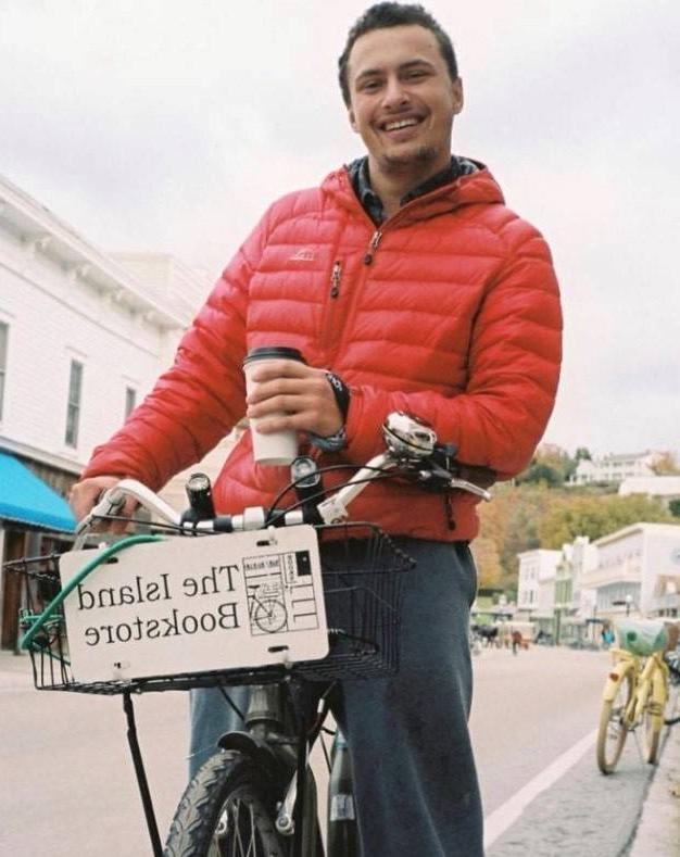 Will Barnwell smiling on a bicycle and holding a cup of coffee